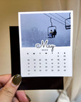 A person's hand holding a small calendar card with a photo of a ski lift and the word "May" written on it.