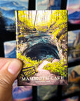A hand holding a magnet depicting the Mammoth Cave National Park, with a scenic view of the cave entrance surrounded by lush vegetation.