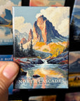 A hand holding a magnet with a painting of a mountain landscape, specifically depicting the North Cascades National Park.