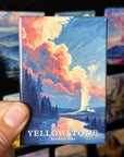 A hand holding a magnet with a colorful illustration of a landscape, depicting a scenic view of Yellowstone National Park.