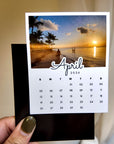 A person's hand holding a calendar for the month of April, with a beach scene visible on the cover.
