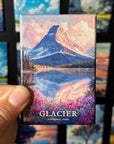 A hand holding a magnet with a painting of a mountain landscape, featuring a lake and colorful flowers.