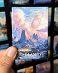 A hand holding a magnet with a mountain landscape and clouds on it, with the text "BADLANDS NATIONAL PARK" visible.