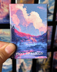 A hand holding a magnet with a colorful illustration of Hot Springs National Park, featuring a scenic landscape with a waterfall and clouds.