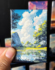 A hand holding a magnet with a painting of a landscape featuring a lake, trees, and a cloudy sky.
