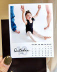 A person is holding a small calendar with a photo of a young girl in a black outfit, smiling and raising her arms.