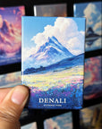 A hand holding a magnet with a mountain landscape and clouds on it, with the word "DENALI" written below the image.