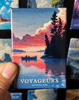 A hand is holding a magnet with a colorful illustration of a boat on a lake, surrounded by trees and a sunset sky.