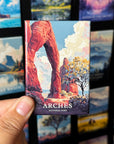 A hand holding a magnet with an illustration of a red rock formation, which appears to be the Arches National Park.