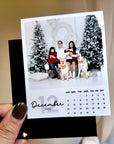 A hand holding a small black and white photo frame with a family portrait of a man, woman, and two dogs, along with a calendar for the month of December 2020.