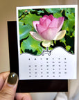 A hand holding a small calendar with a pink flower image on the cover.