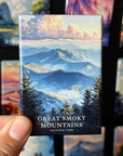 A hand holding a magnet with a scenic mountain landscape featuring clouds and pine trees.