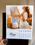 A hand holding a small calendar with a photo of a couple on the cover, and a calendar for the month of May.