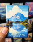 A hand is holding a magnet with a painting of Lake Clark and its surroundings, including a mountain and a lake, against a background of other landscape paintings.