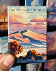 A hand holding a magnet with an illustration of sand dunes and mountains, with the text "GREAT SAND DUNES NATIONAL PARK" printed on it.