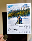 A person is holding a small calendar with a photo of a couple embracing in front of a snowy mountain landscape.
