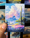 A hand holding a magnet with a colorful illustration of a desert landscape, including mountains, cacti, and flowers.