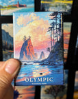 A hand holding a magnet with a scenic landscape of a mountain and a body of water, with the text "OLYMPIC NATIONAL PARK" visible.