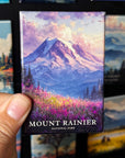 A hand holding a magnet with a painting of a mountain landscape, specifically Mount Rainier, in the background.