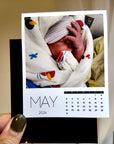 A person's hand holding a small, square-shaped calendar with a photo of a baby wrapped in a blanket on the cover.