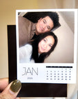 A person is holding a small square photo frame containing a photo of a man and a woman, along with a calendar for the month of January.