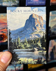 A hand holding a magnet with a picture of Rocky Mountain National Park, featuring a mountain landscape with a lake and forest in the foreground.
