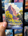 A hand holding a magnet with a painting of a mountain landscape, specifically Capitol Reef National Park, against a background of other landscape paintings.