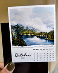 A hand holding a calendar for October 2014, featuring a scenic landscape with a lake and mountains.