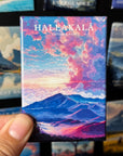 A hand holding a magnet with a colorful landscape depicting a mountain range and a sunset sky.
