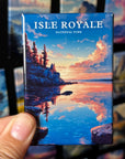 A hand holding a magnet with a scenic image of Isle Royale National Park, featuring a sunset over the water and rocky cliffs.