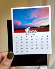 A person's hand holding a small calendar card with a beach scene and the word "June" written on it.