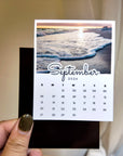 A person's hand holding a calendar card with a beach scene on the cover.