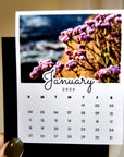 A person's hand holding a calendar card with a photo of purple flowers on the cover.