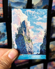 A hand holding a magnet with a scenic mountain landscape, including a river and clouds, against a background of other magnets.