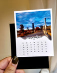 A person's hand holding a small calendar card with a photo of a city street and buildings.