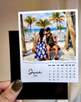 A person is holding a small photo frame containing a printed photo of a couple sitting on a swing, with a beach scene in the background.