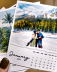 A hand holding a calendar with a photo of a couple in front of a snowy mountain landscape.