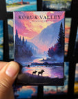 A hand holding a magnet with a scenic landscape of Kobuk Valley National Park, featuring silhouettes of moose and trees against a colorful sunset sky.
