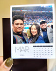 A person is holding a small photo frame with a photo of three people inside, and the frame has a calendar for the month of March.