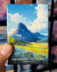A hand holding a magnet with a painting of a mountain landscape, specifically Wrangell-St. Elias National Park.
