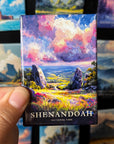 A hand holding a magnet with a colorful painting of a scenic landscape, featuring mountains, clouds, and a river.