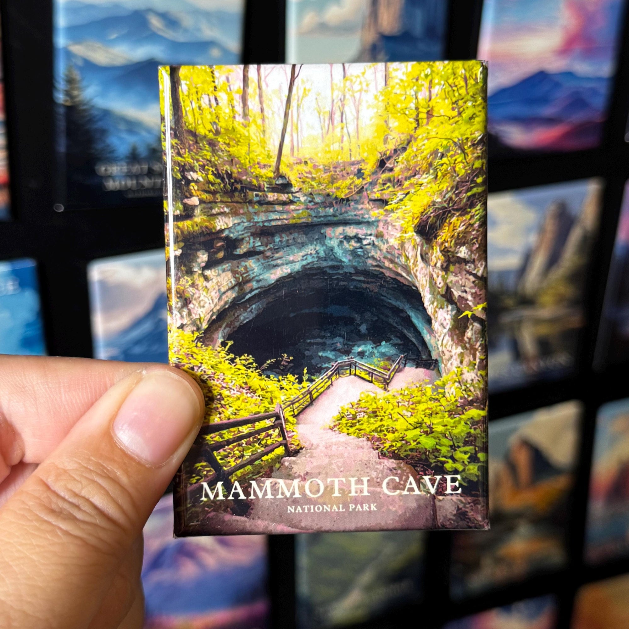 A hand holding a magnet depicting the Mammoth Cave National Park, with a scenic view of the cave entrance surrounded by lush vegetation.