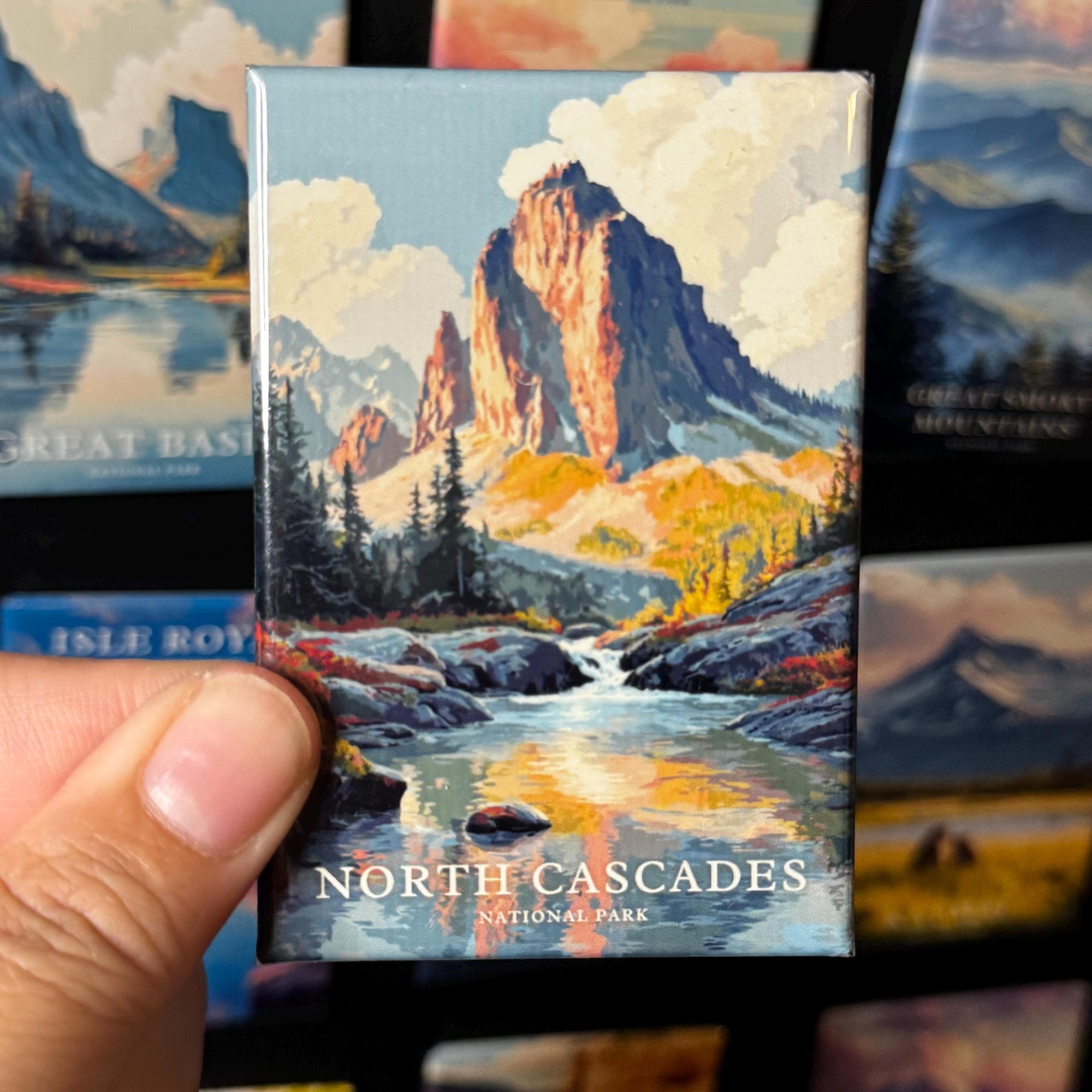 A hand holding a magnet with a painting of a mountain landscape, specifically depicting the North Cascades National Park.