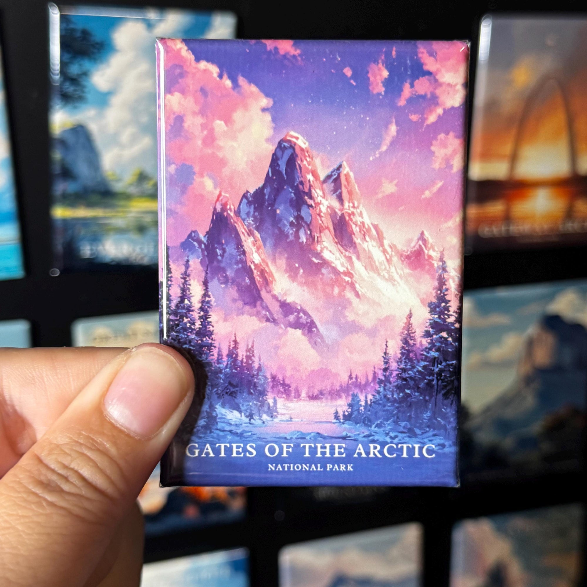 A hand holding a magnet with a colorful illustration of a mountain landscape, with the text "GATES OF THE ARCTIC NATIONAL PARK" printed on it.