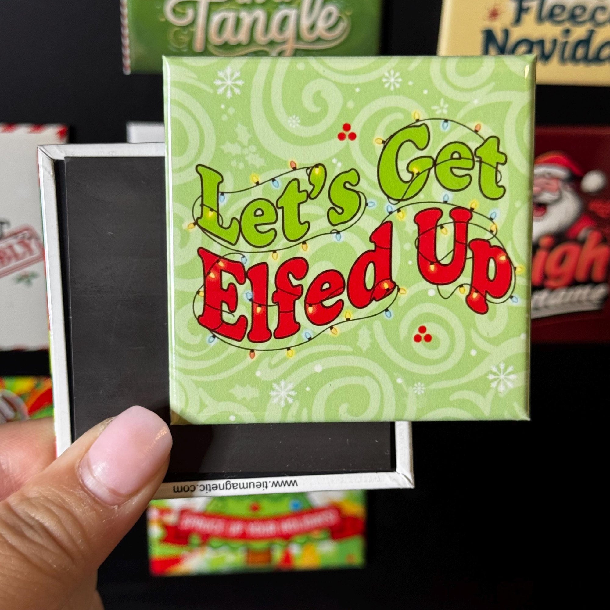 A hand holding a green magnet with the text "Let's Get Elfed Up" on it, which appears to be a humorous Christmas-themed magnet.