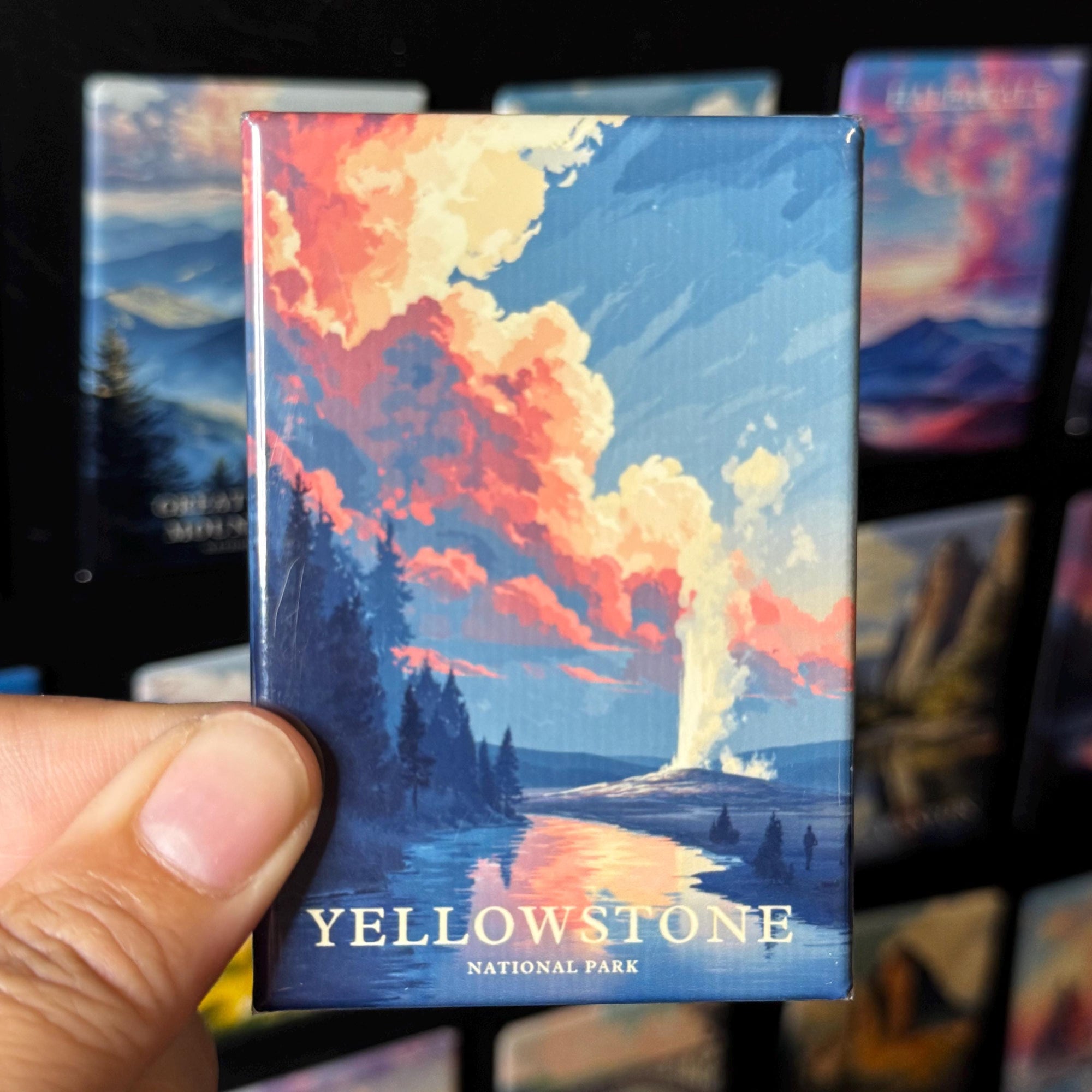 A hand holding a magnet with a colorful illustration of a landscape, depicting a scenic view of Yellowstone National Park.