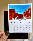A person's hand holding a calendar card with a photo of a red rock formation and the word "November" written on it.