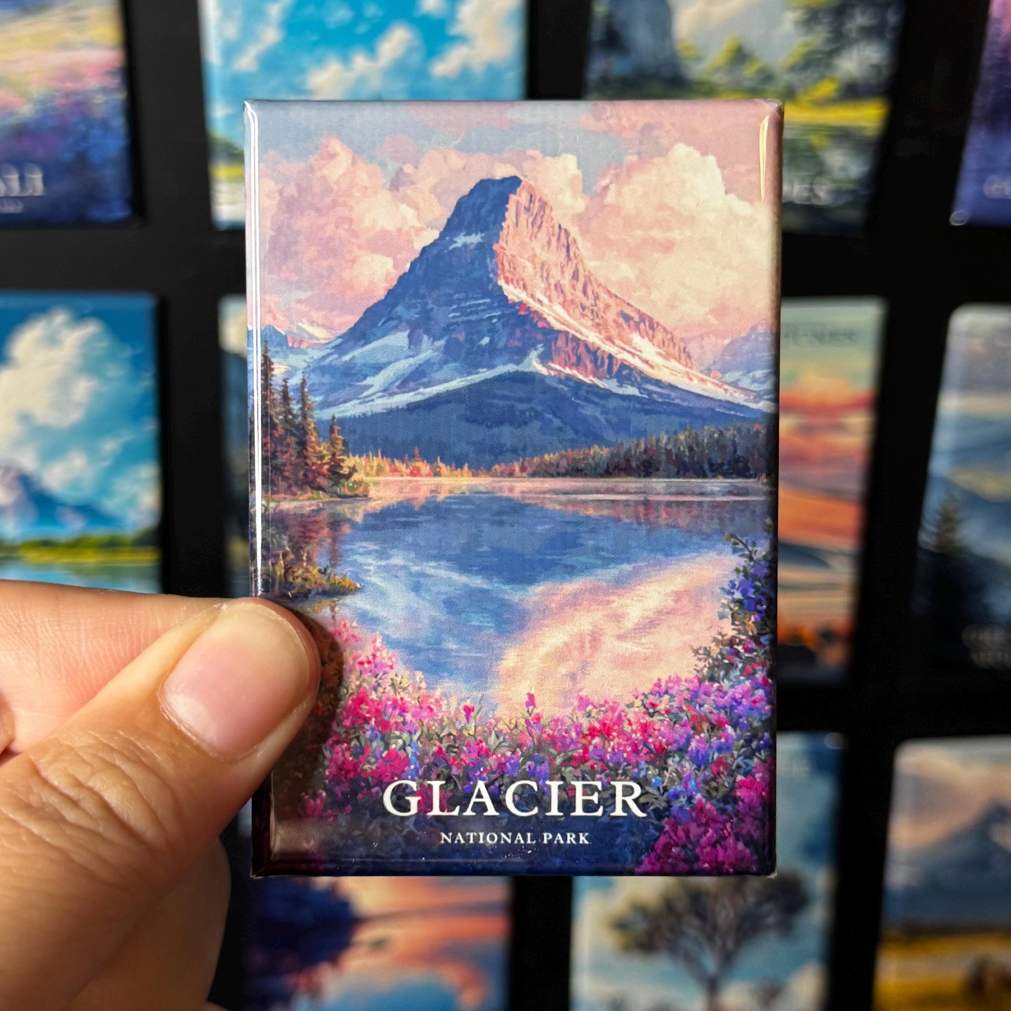 A hand holding a magnet with a painting of a mountain landscape, featuring a lake and colorful flowers.
