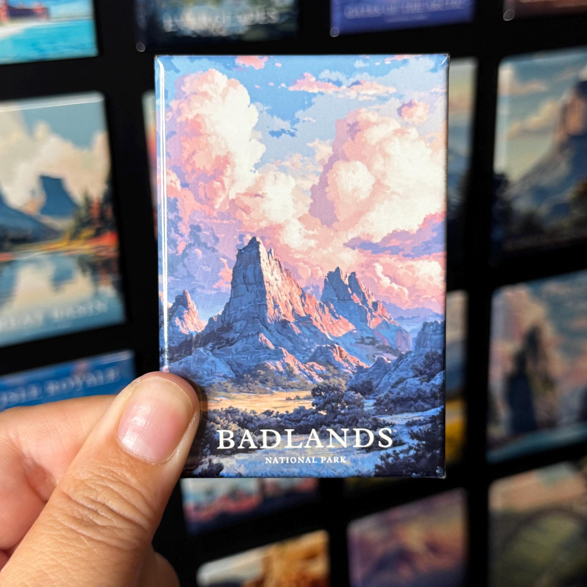 A hand holding a magnet with a mountain landscape and clouds on it, with the text "BADLANDS NATIONAL PARK" visible.