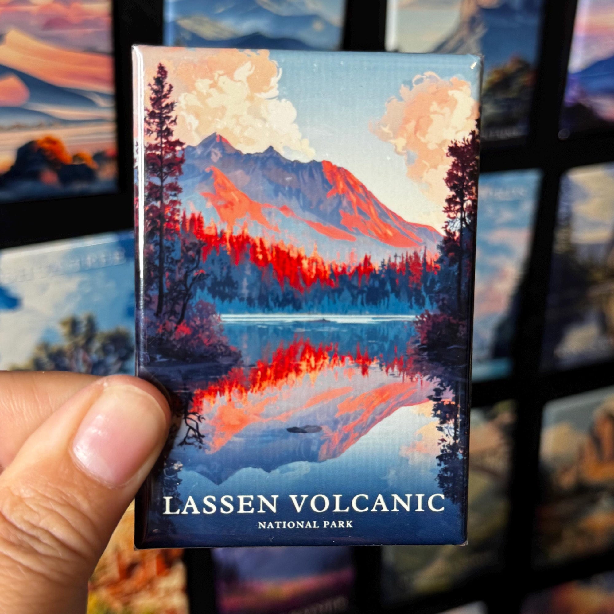 A hand holding a magnet with a landscape depicting a mountain and a lake, with the text "LASSEN VOLCANIC NATIONAL PARK".